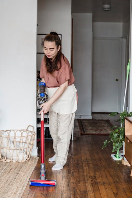 Two professional women in beige cleaning uniforms standing against a plain white wall, holding cleaning tools—a vacuum cleaner and a mop—ready for domestic cleaning tasks. Their uniforms are clean
