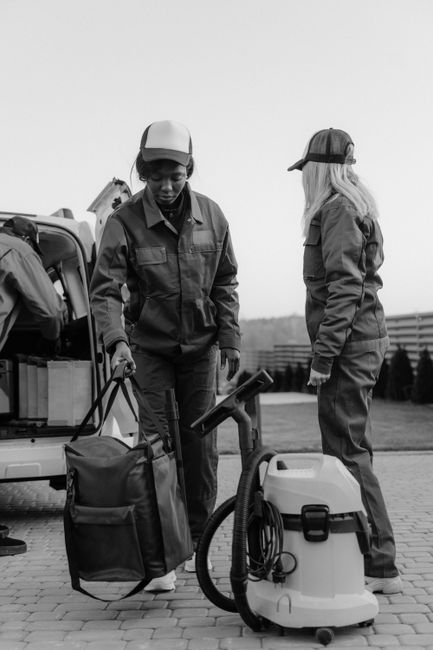 Two professional cleaners from West Kensington Cleaners are preparing for a deep cleaning session outdoors on a paved area. One cleaner, wearing a jacket and cap, is examining a clipboard while standi