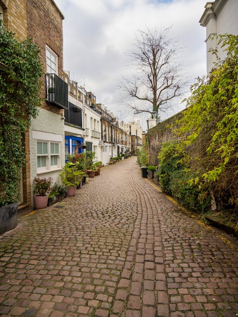 A narrow cobblestone street in West Kensington, lined with residential buildings featuring white and brick façades. The street is bordered by potted plants and greenery along the base of the building