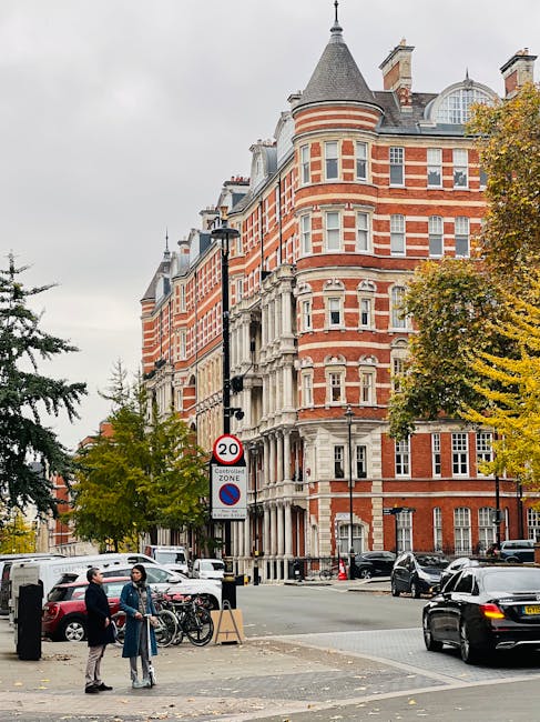A narrow cobblestone street in West Kensington, lined with residential buildings featuring white and brick façades. The street is bordered by potted plants and greenery along the base of the building