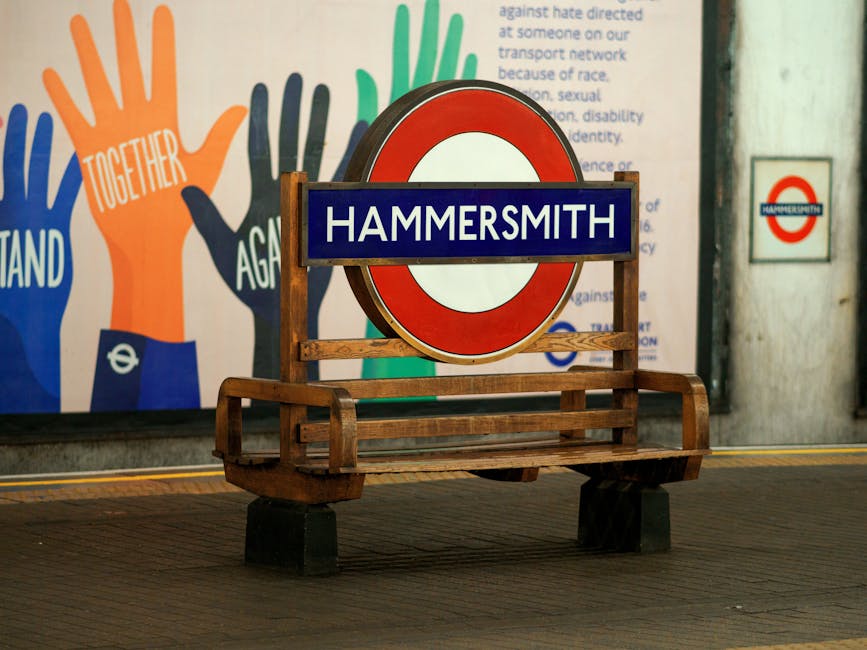 A wooden bench with a backrest and armrests, situated on a tiled platform at Hammersmith underground station in London. Behind the bench, there is a large round London Underground sign with a blue rec